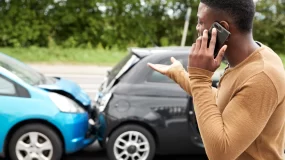 Upset African American man on phone looking at car wreck.