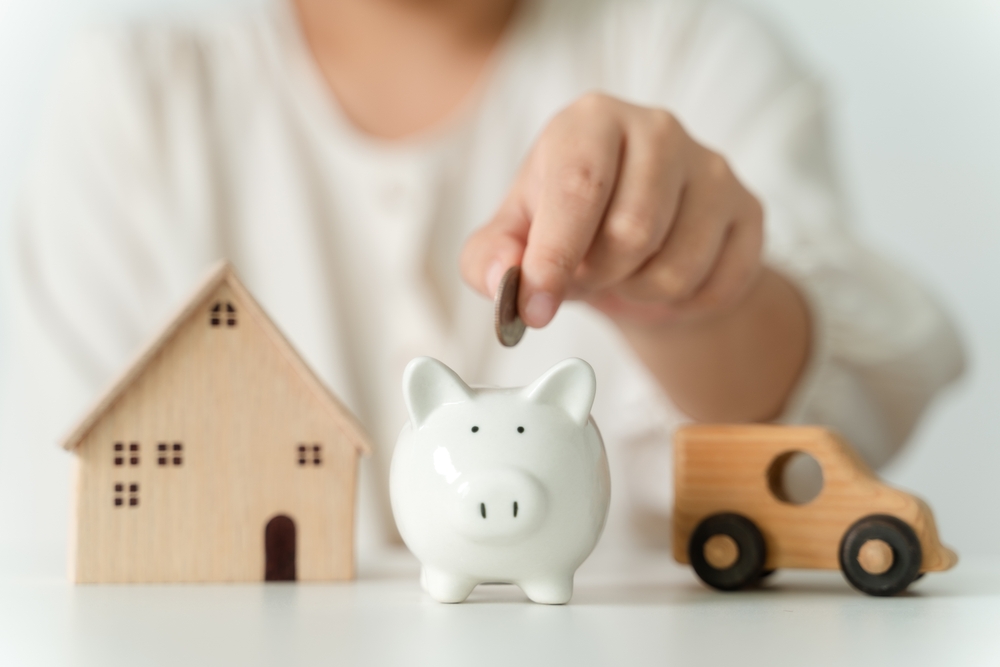 Hand putting a coin into a miniature piggy bank with a wooden car and house in the background, symbolizing bundling - car insurance in Georgia